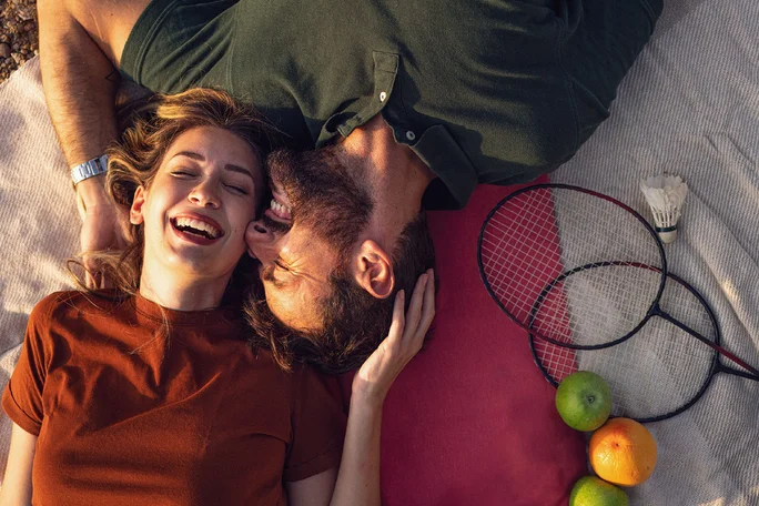 Par leži na dekici na otvorenom, kraj njih su reketi za badminton, loptica i voće. Muškarac se igra ljubeći nasmiješenu ženu u obraz.