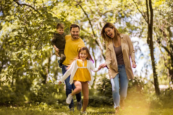 A family of four walking outdoors in a sunlit forest or park, holding hands and smiling. The father, wearing a yellow shirt and jeans, carries a young boy dressed in dark clothing. The mother, in a beige jacket and jeans, holds the hand of a young girl wearing a yellow dress and white cardigan. The background is filled with green trees and sunlight filtering through the leaves.