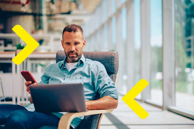 Muškarac sjedi i radi nešto na laptopi i drži mobitel u ruci