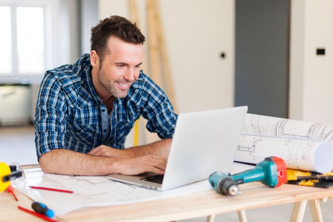 smiling man working on laptop surrounded with construction tools nearby